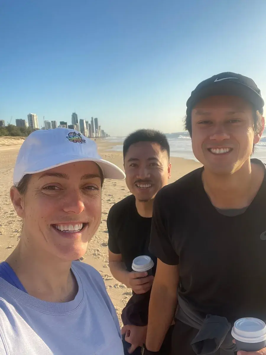 Three teammates on a morning beach walk, coffees in hand, with the city skyline behind