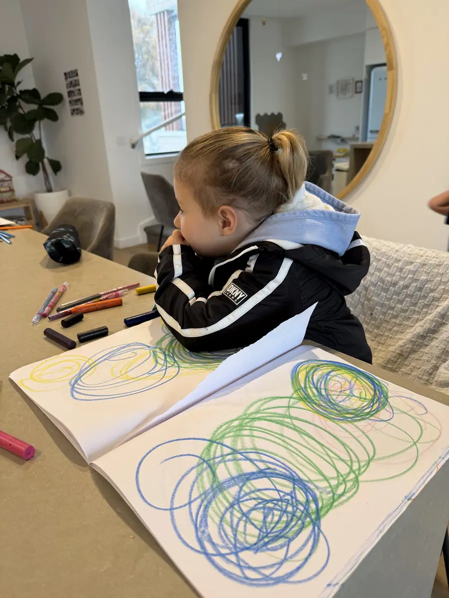 A child resting her chin on her arms at the kitchen bench, beside pages of looping crayon circles